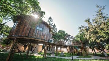 Una casa en el árbol de madera con una plataforma circular de observación, sostenida por vigas de madera, en un parque frondoso.