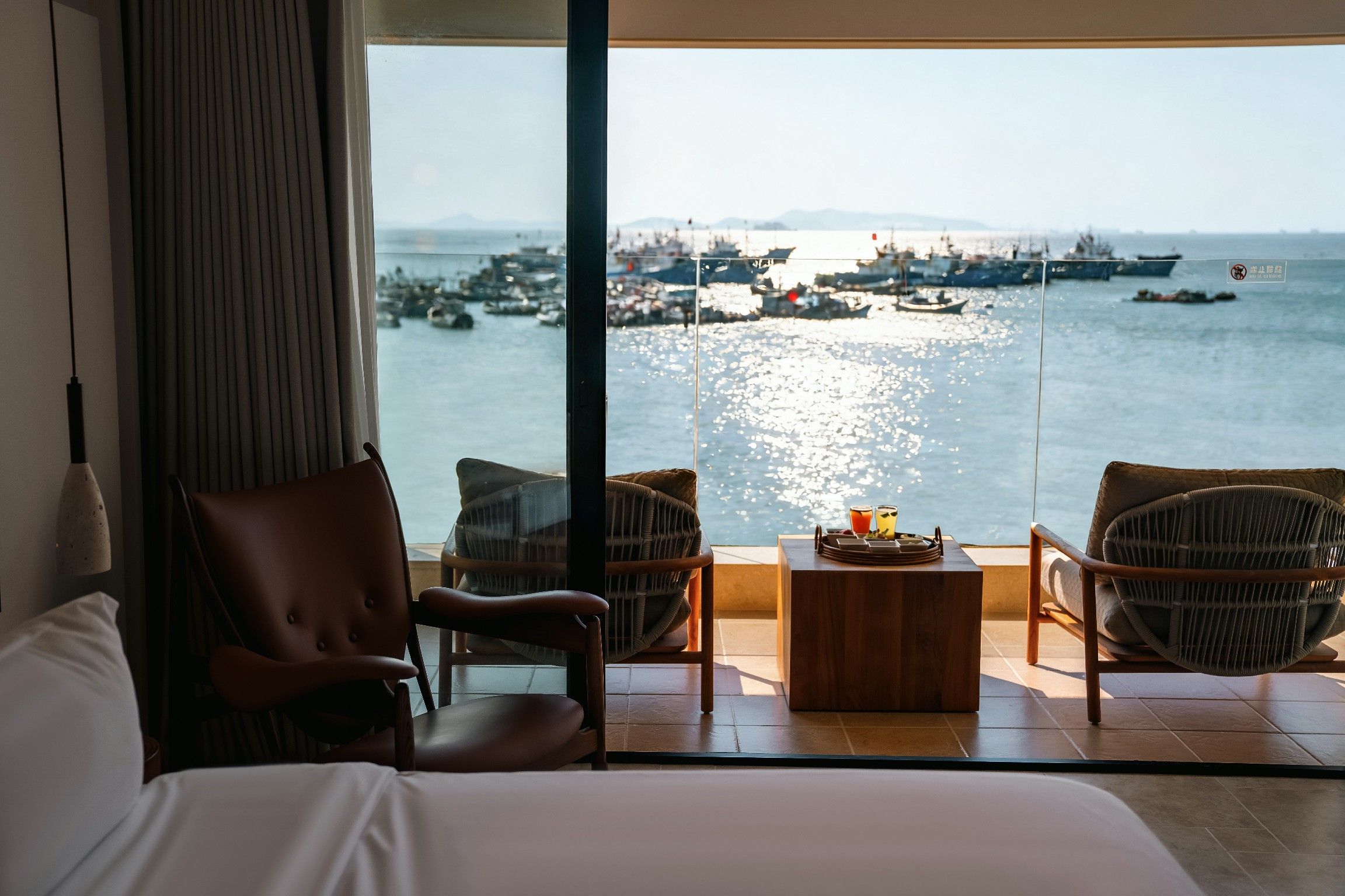 Habitación de hotel con un ventanal grande que ofrece vistas al mar y barcos. Incluye una cama, dos sillas de mimbre y una pequeña mesa de madera con bebidas.