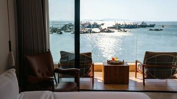 Habitación de hotel con un ventanal grande que ofrece vistas al mar y barcos. Incluye una cama, dos sillas de mimbre y una pequeña mesa de madera con bebidas.