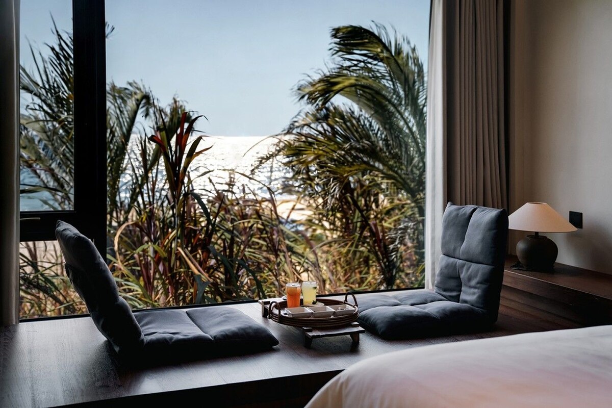 A hotel room with a bed, two chairs, a small table with drinks, and a view of palm trees.