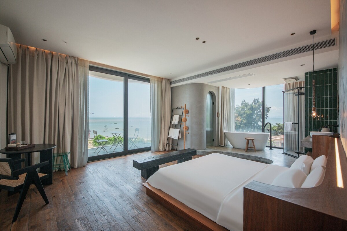 A modern bedroom with a large bed, a sitting area, and a view of the sea.