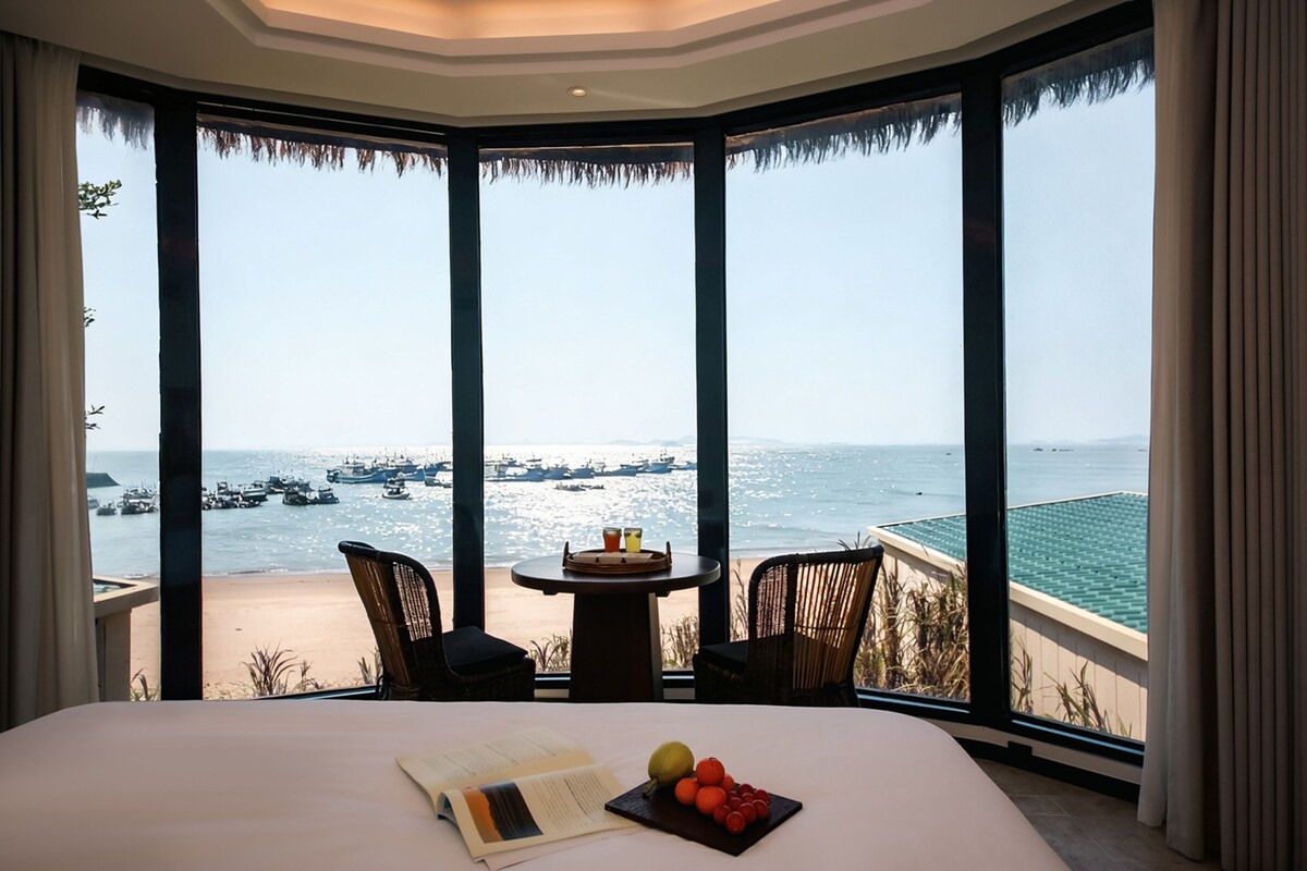 A hotel room with a large bed, a small table with drinks, and a view of the sea with boats.