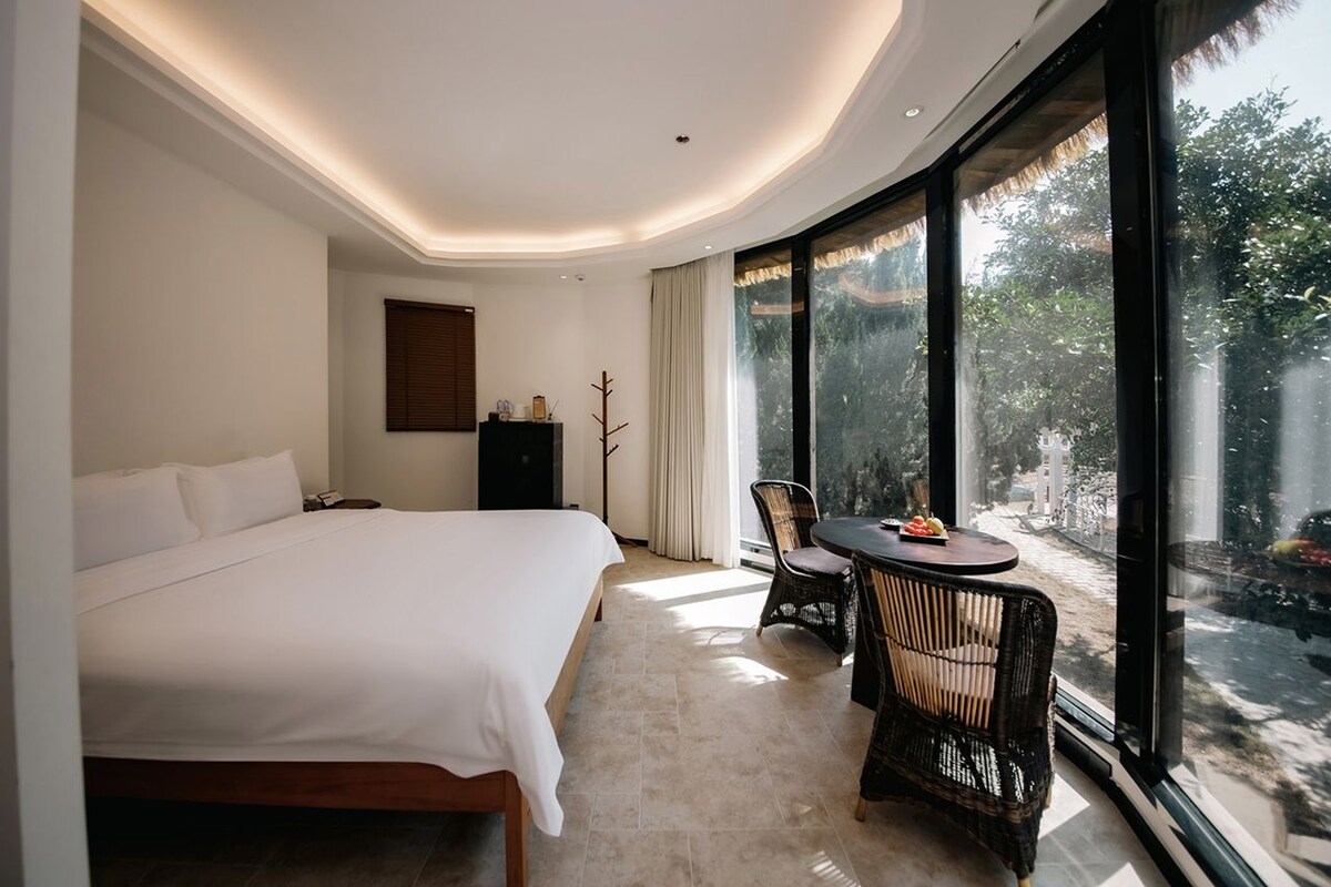 A hotel room with a large bed, a dining table with chairs, and a view of the outdoors through large windows.