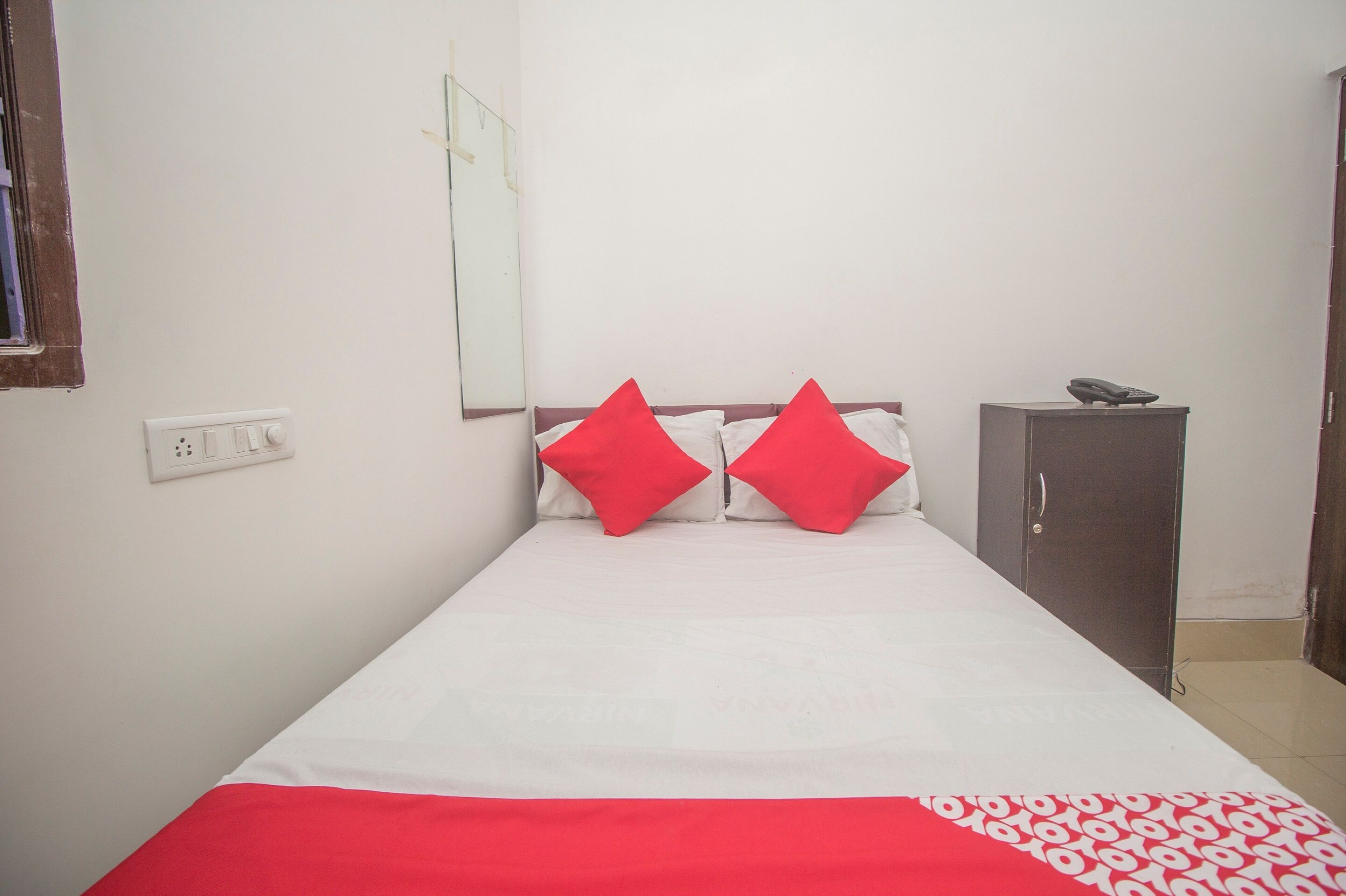 A single bed with a white mattress and red pillows, a small bedside table with a phone, and a cabinet against the wall.