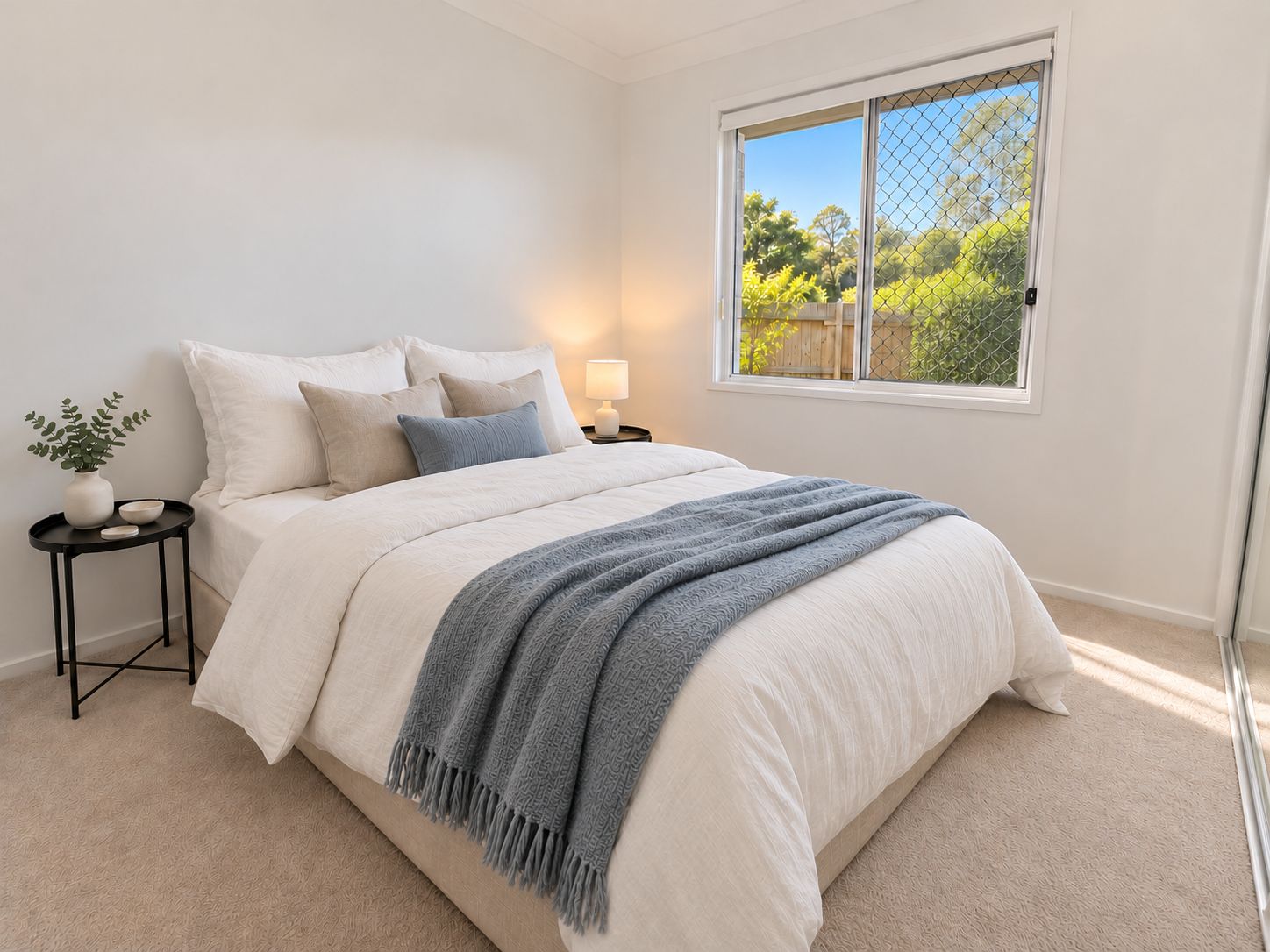 A bedroom with a bed, a bedside table, a lamp, a window, and a view of greenery outside.