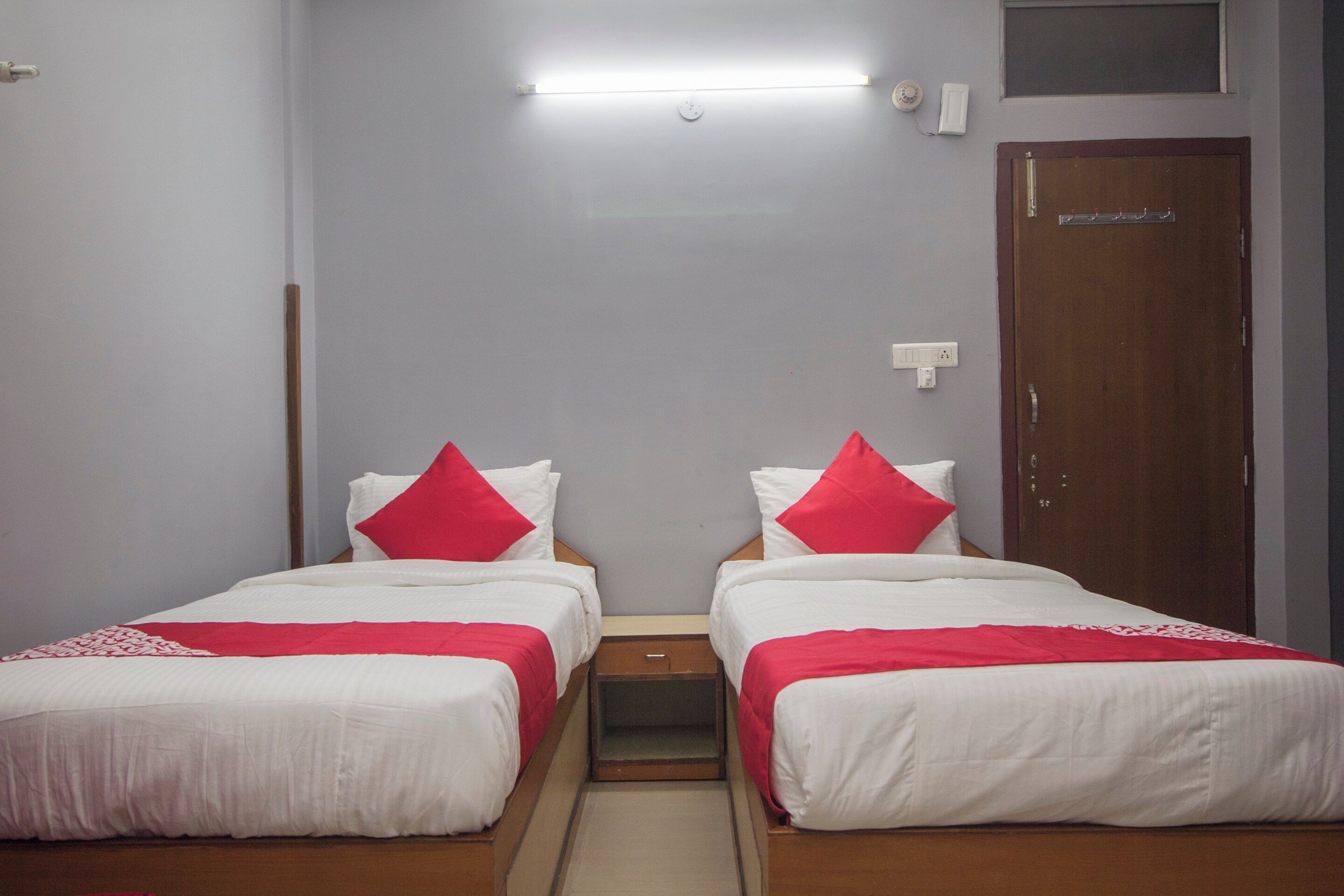 A hotel room with two single beds, each with white bedding and red pillows, a small bedside table, a wooden wardrobe, and a light fixture on the ceiling.