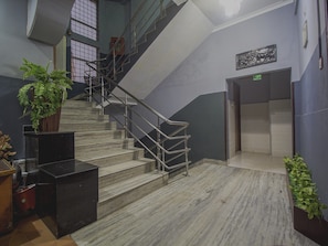 A staircase with metal railings leading to an elevator, surrounded by walls with a geometric pattern and a potted plant on the left. - Hotel O Golden Moments (Siliguri)