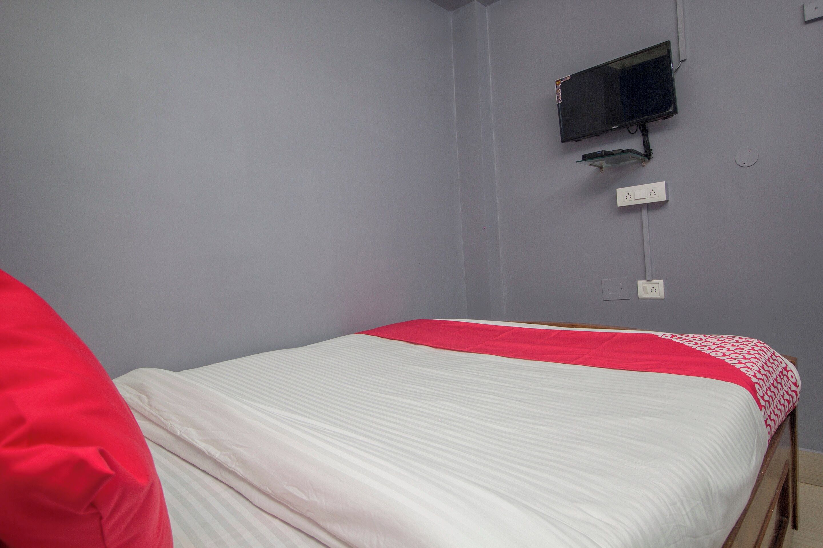 A single bed with a white and red bedspread, a television mounted on the wall, and a power outlet below it.