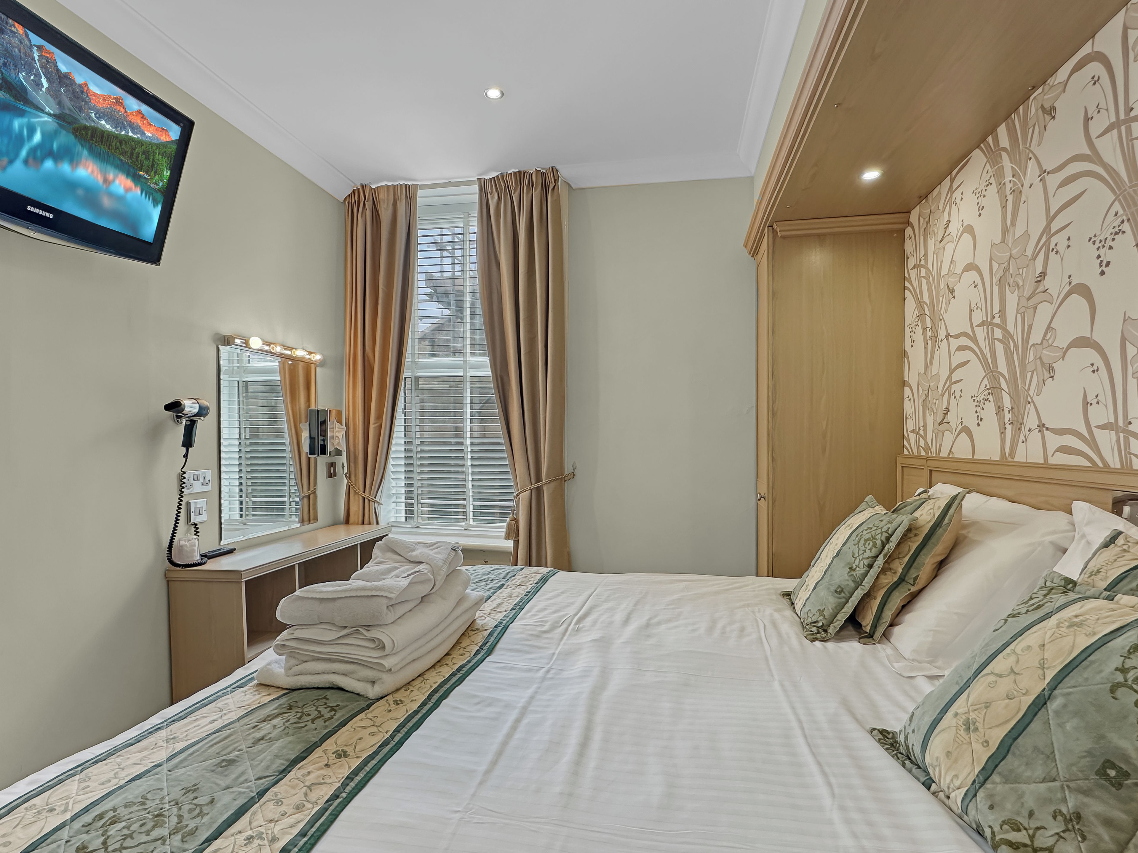 A hotel room with a bed, a vanity with a mirror, a television mounted on the wall, and a window with curtains.