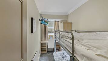 Une chambre d’hôtel étroite, avec un lit superposé, un téléviseur fixé au mur, un radiateur et une fenêtre avec des rideaux.