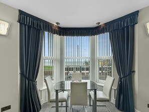 A dining area with a round table and two chairs, large windows with vertical blinds, and a view of a building with a colorful facade. - Granada Apartments Burbage flat 4 1st floor sea view (Blackpool FY2 9JS)