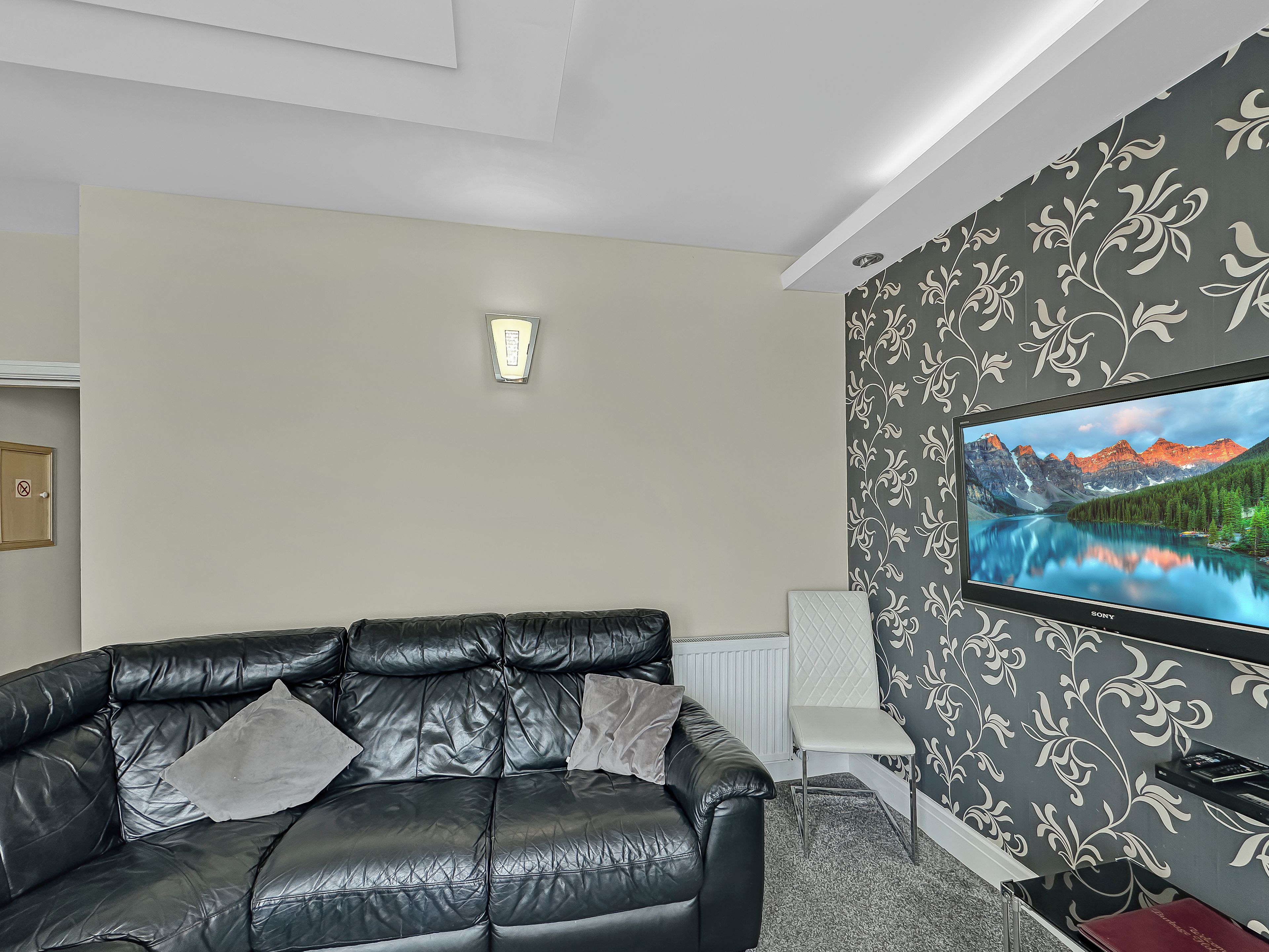 A living room with a black leather sofa, a flat-screen TV, and a patterned wallpaper.