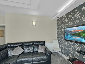 A living room with a black leather sofa, a flat-screen TV, and a patterned wallpaper. - Granada Apartments Burbage flat 4 1st floor sea view (Blackpool FY2 9JS)
