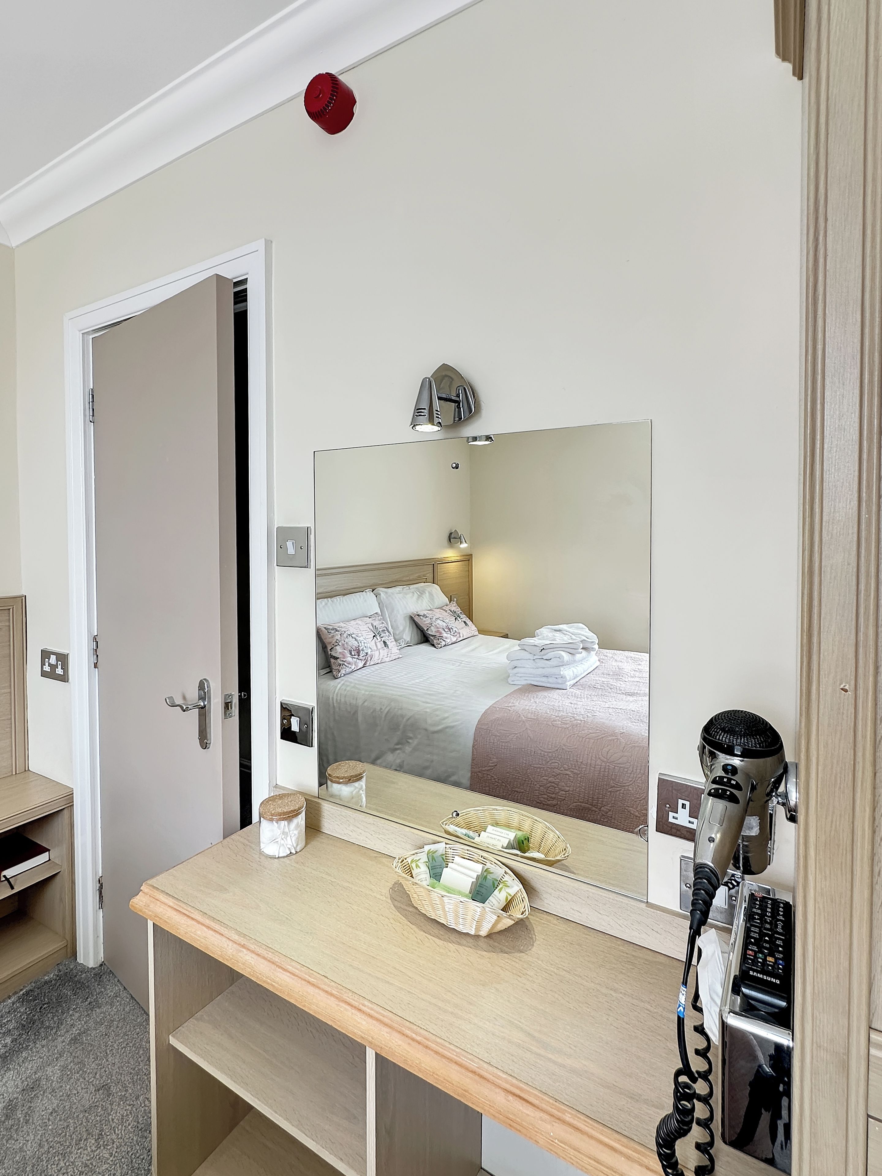 A hotel room with a wooden vanity, a mirror, a hairdryer, a telephone, and a bed with a headboard.