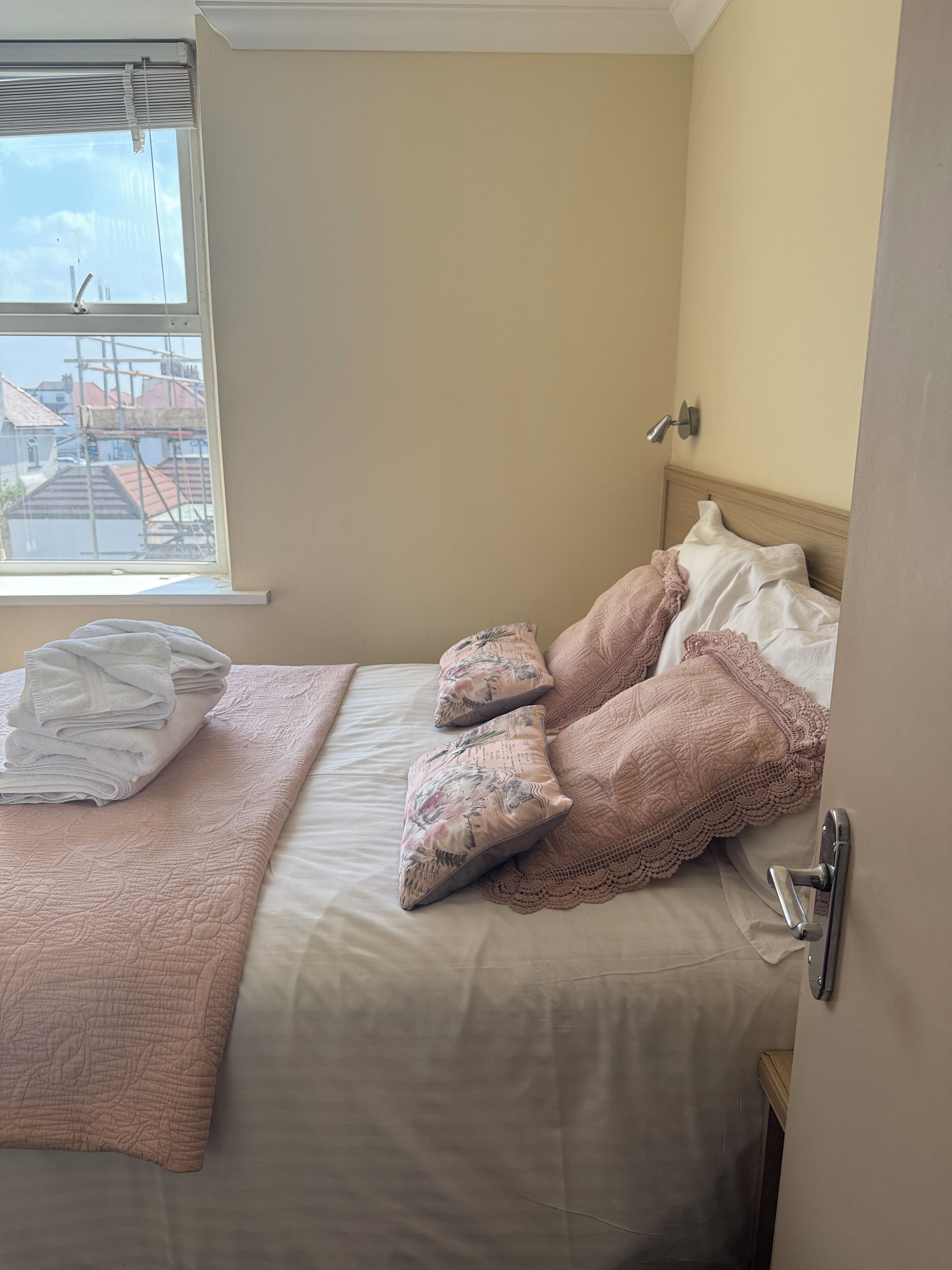 A hotel room with a bed, pillows, a folded towel, a bedside lamp, and a window with a view of buildings.