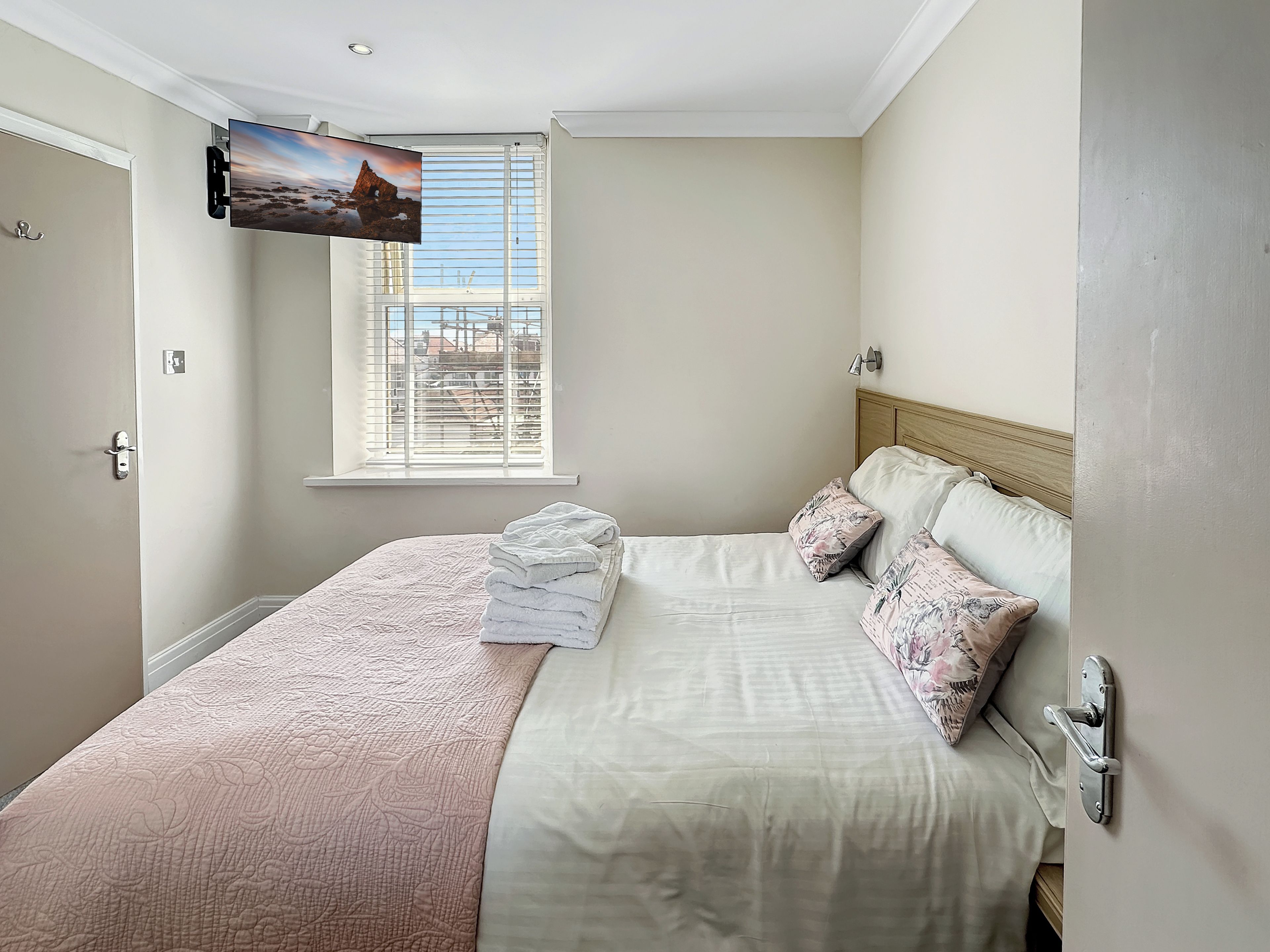A bedroom with a bed, a TV mounted on the wall, a window with blinds, and a door.