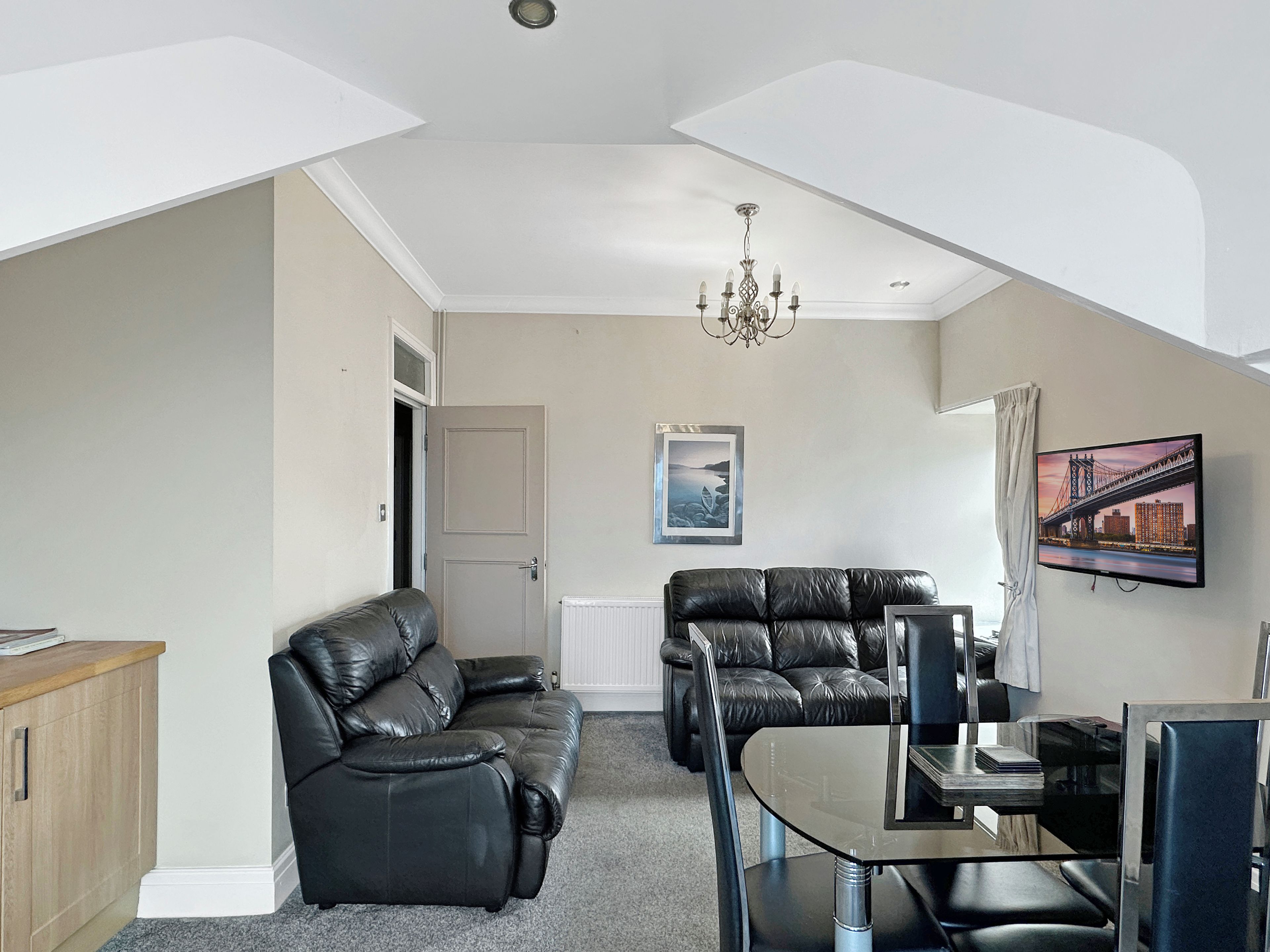 A modern living room with a high ceiling, a chandelier, a flat-screen TV mounted on the wall, and a dining area with a glass table.