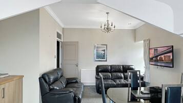 A modern living room with a high ceiling, a chandelier, a flat-screen TV mounted on the wall, and a dining area with a glass table.