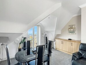 A modern loft apartment with a dining area, a black leather sofa, a wooden cabinet, and a view of a cityscape through a large window. - Granada Apartments Burbage flat 6 2nd floor front sea view (Blackpool FY2 9JS)