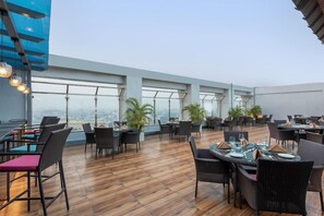 An outdoor dining area with tables and chairs, large windows, and potted plants. - Park Inn by Radisson Lucknow Vikas Nagar (Unnao)