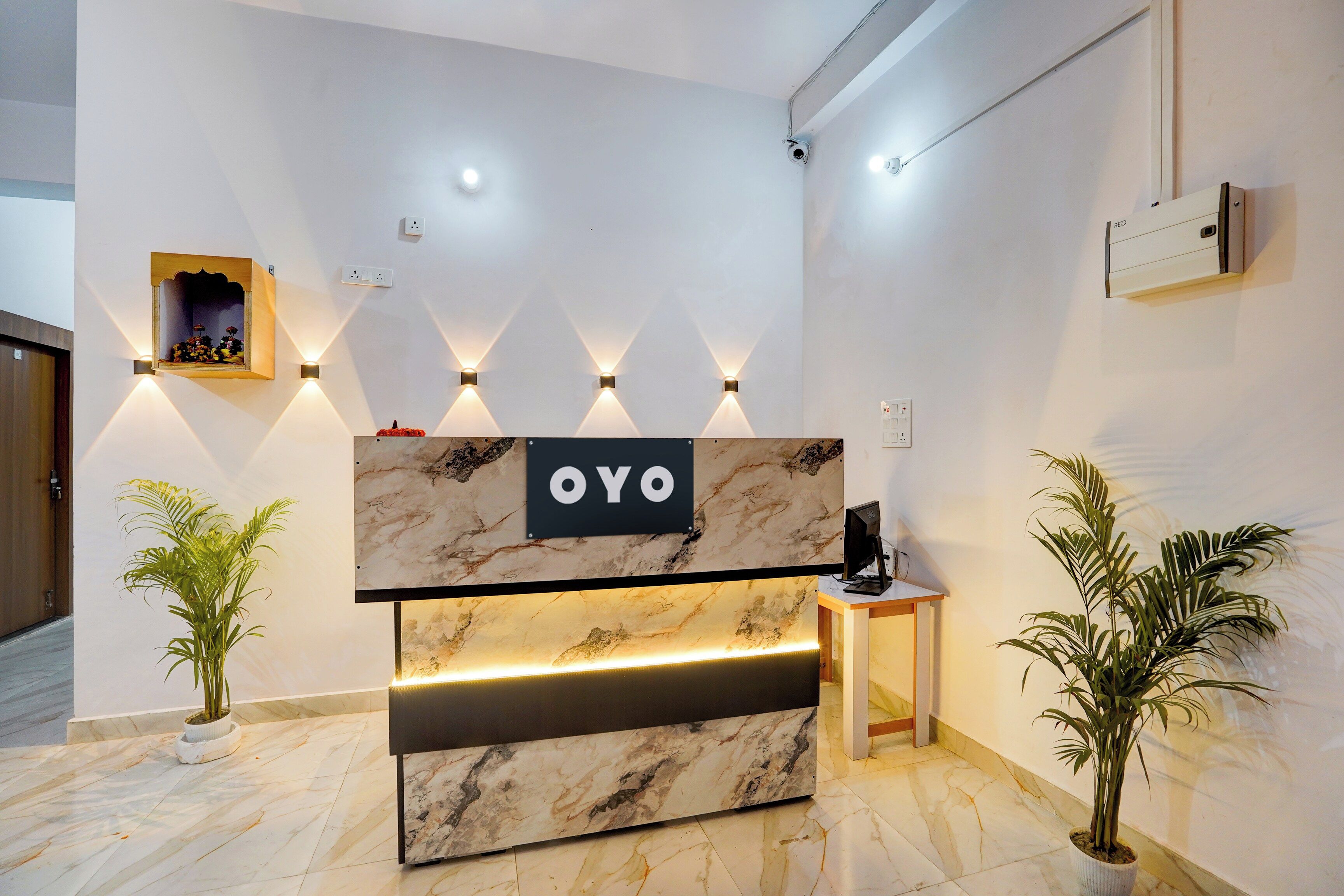 Tauleller de recepció amb superfície de marbre i un cartell que diu 'OYO'. Hi ha llums penjades a la paret sobre la taula, una petita estant amb una fotografia emmarcada i dues plantes en testos a cada costat.