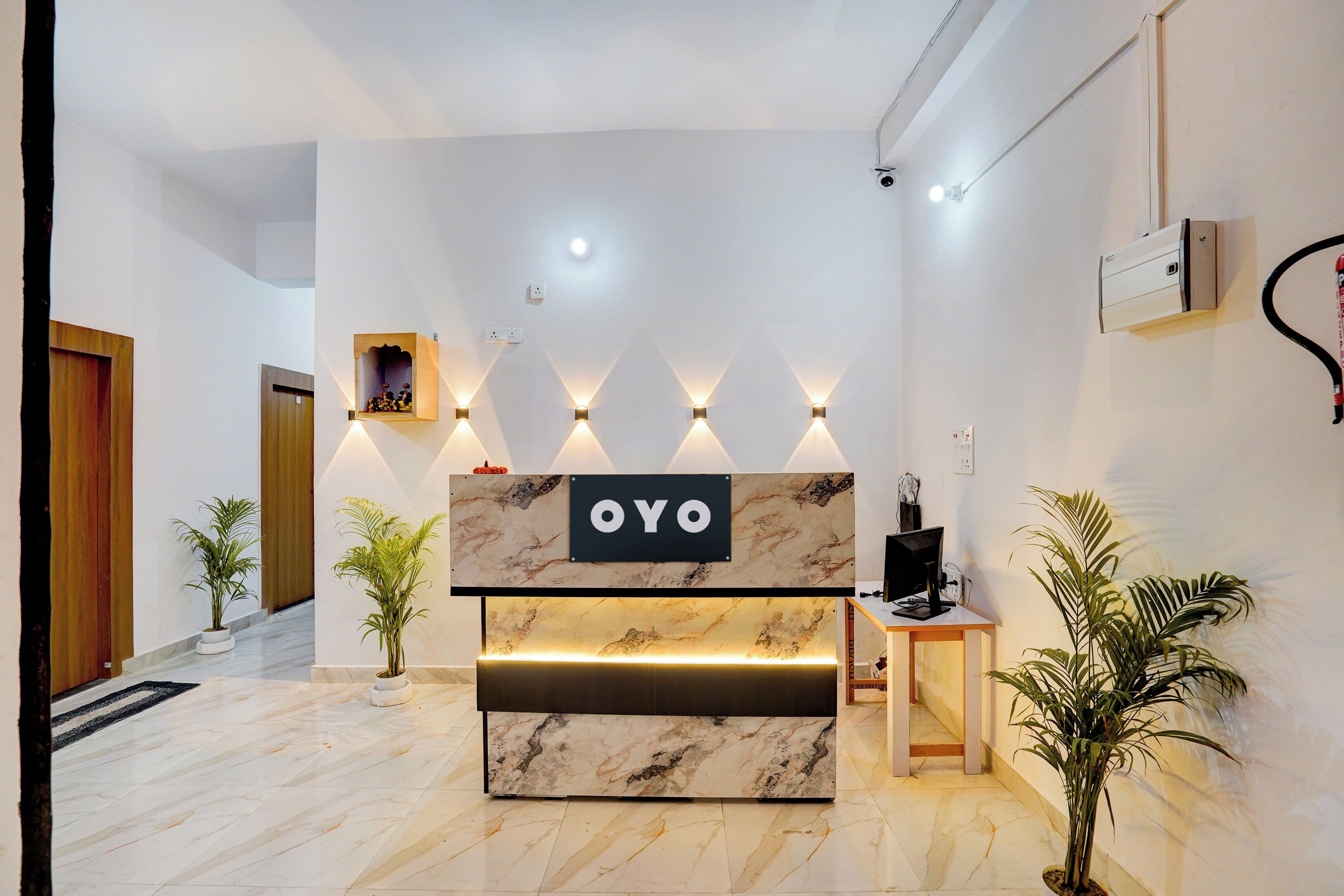 A reception desk with a marble top and a sign that reads 'OYO'. There are wall-mounted lights above the desk, a wooden door to the left, and a staircase leading to an upper level.