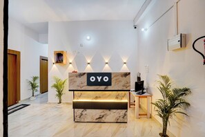A reception desk with a marble top and a sign that reads 'OYO'. There are wall-mounted lights above the desk, a wooden door to the left, and a staircase leading to an upper level. - Hotel O 7 Orchid (Sultanpur)
