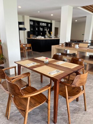 Un comedor con una mesa y sillas de madera, un florero central con una flor, y una barra al fondo.