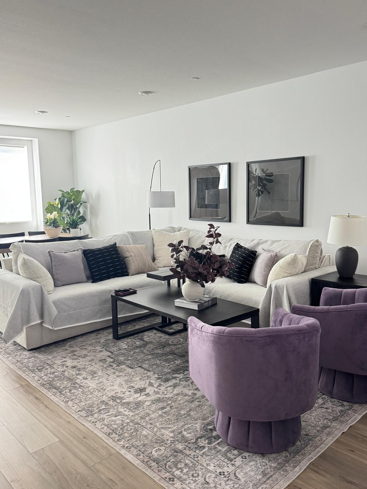 A modern living room with a sofa, coffee table, and two armchairs. There are two framed artworks on the wall and a floor lamp.