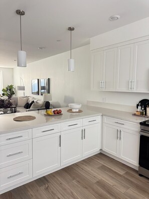 Een moderne keuken met witte kastjes, een centraal eiland, hanglampen en een zithoek met een bank en planten.
