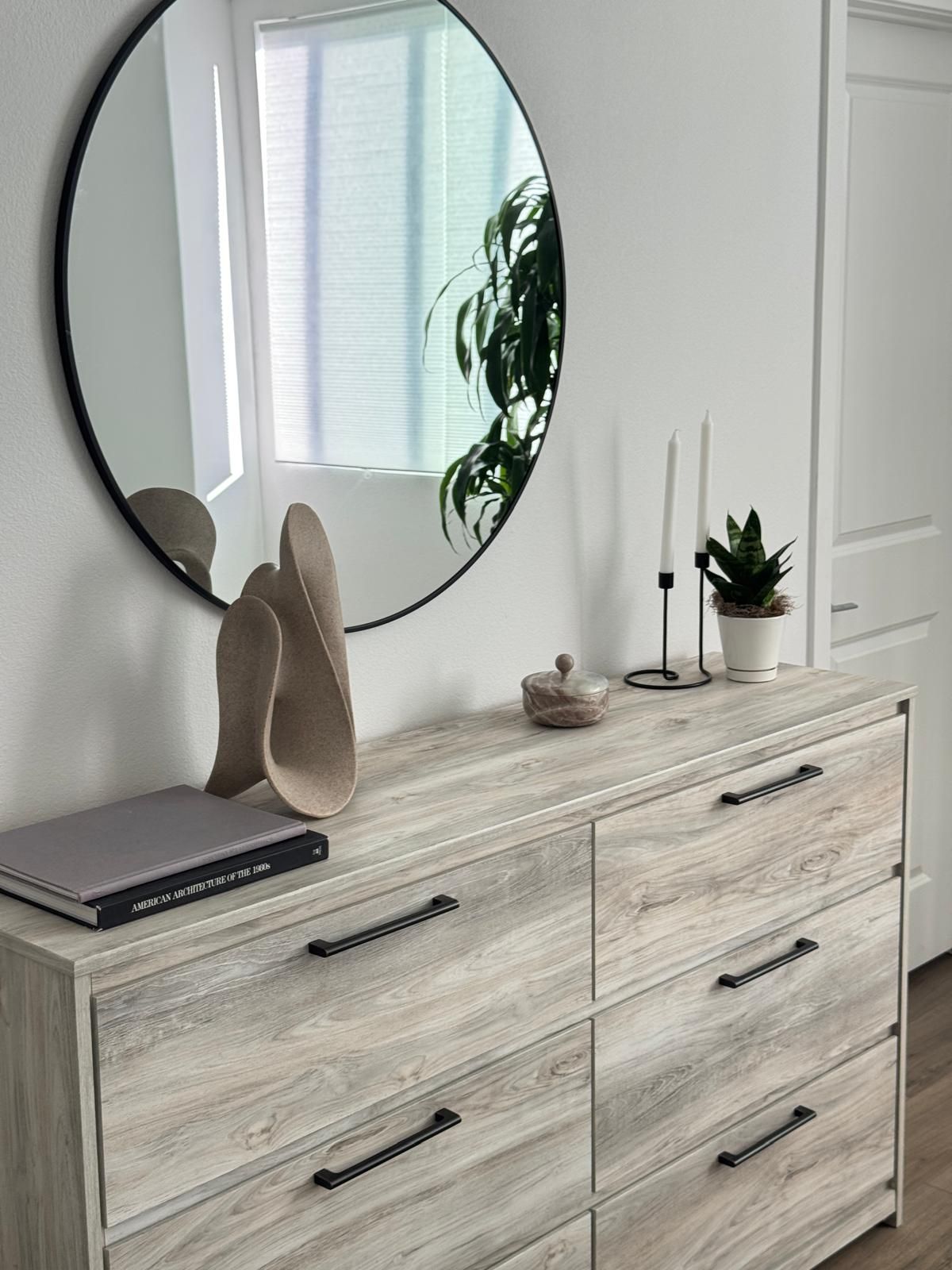 Una stanza moderna e minimalista con un comò in legno chiaro, uno specchio rotondo e una pianta in vaso.
