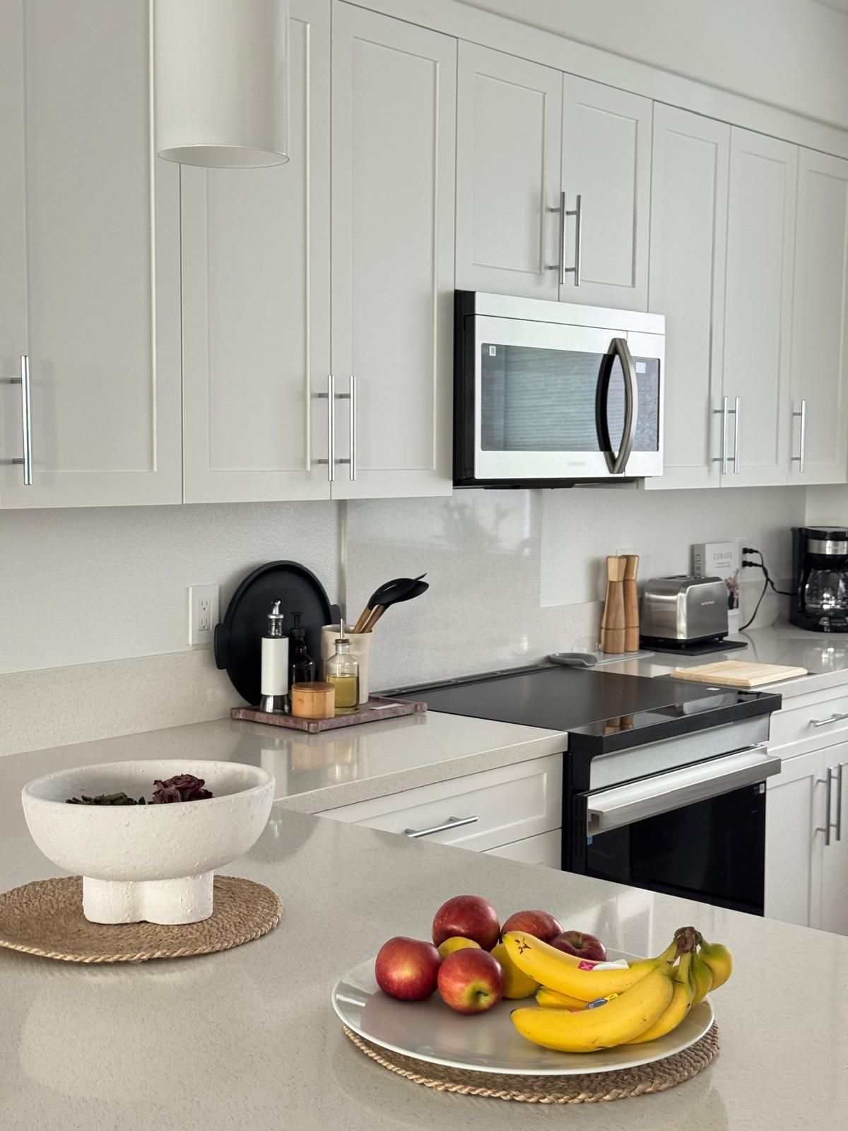 A modern kitchen with white cabinets, a stainless steel microwave, and a countertop with a bowl of fruit.