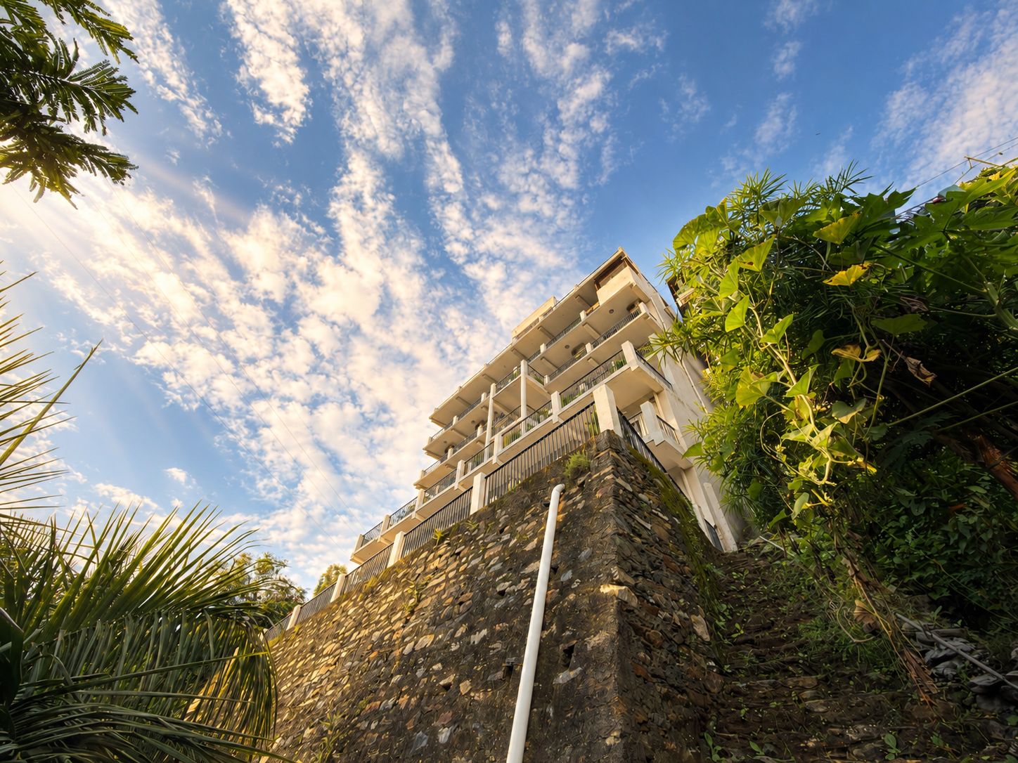 A multi-story building with balconies, surrounded by lush greenery and a clear sky.