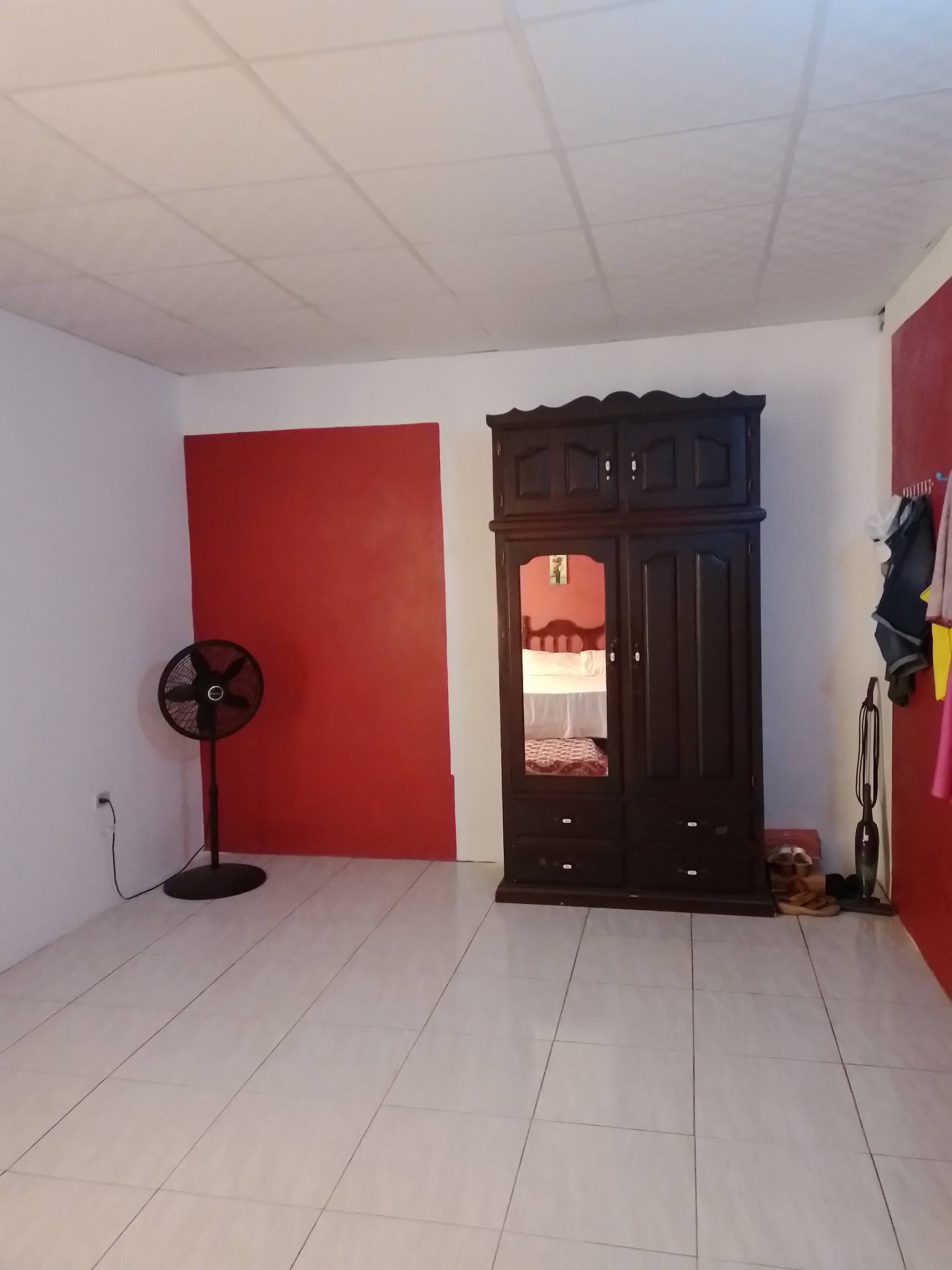 Habitació amb paret vermella, armari de fusta, ventilador de peu i terra emblenquinada.