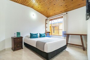 Une chambre avec un plafond en bois, un lit avec une literie blanche et des coussins turquoise, une petite table de chevet en bois et une fenêtre avec des rideaux.