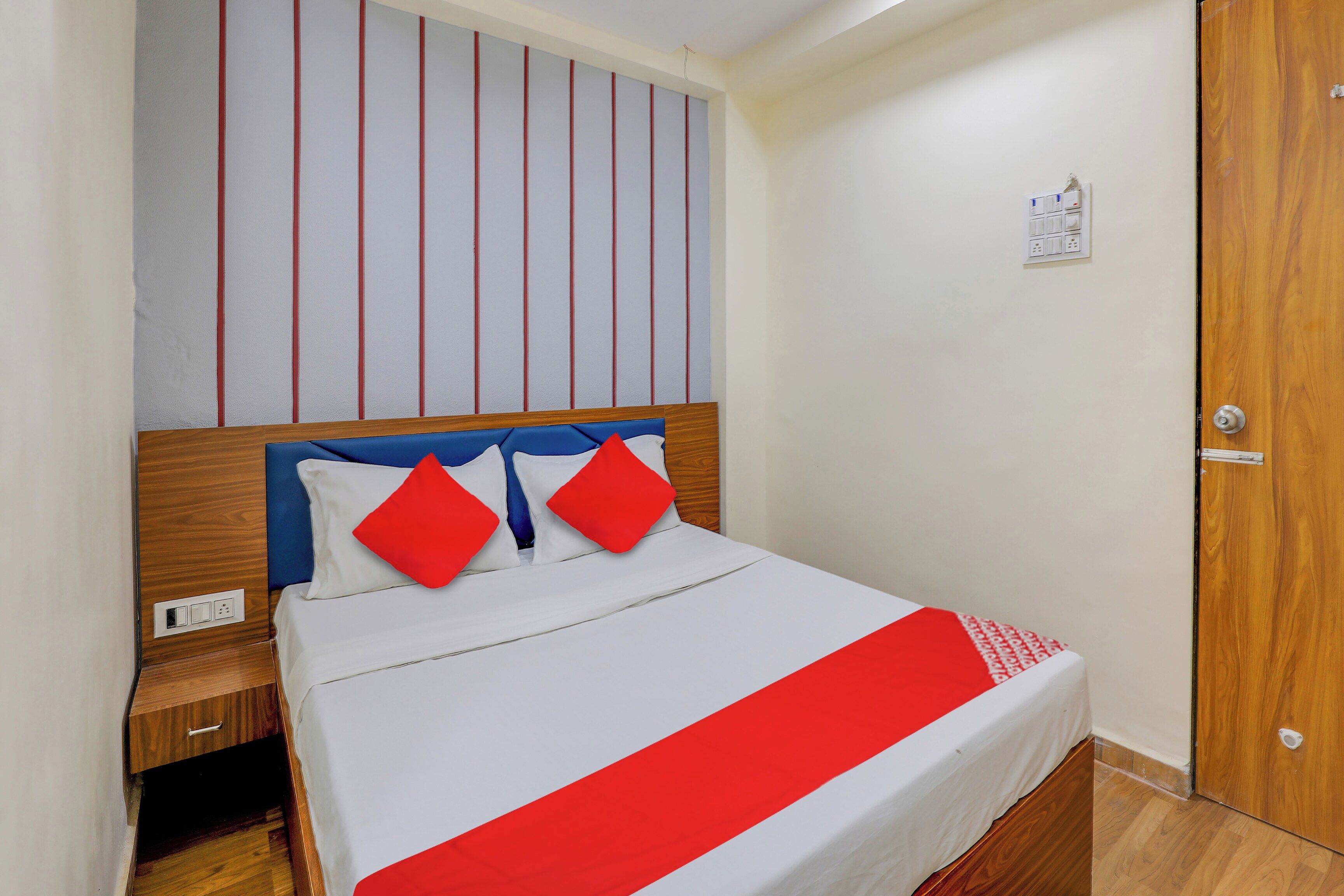 Una habitación de hotel con cama, cabecera de madera, pared a rayas rojas y blancas, y una puerta de madera.