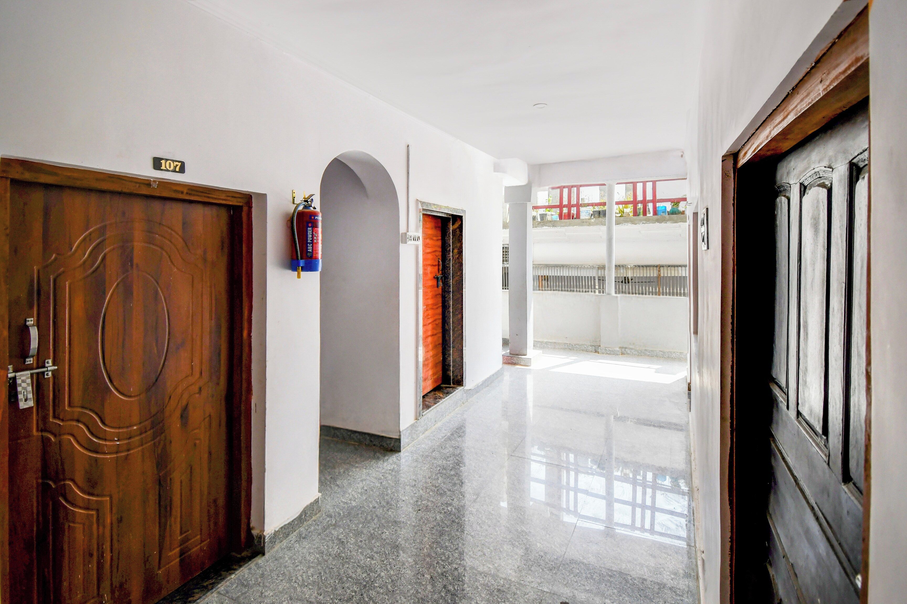 A hallway with a wooden door labeled '107', a fire extinguisher, and a view of a balcony with a railing.