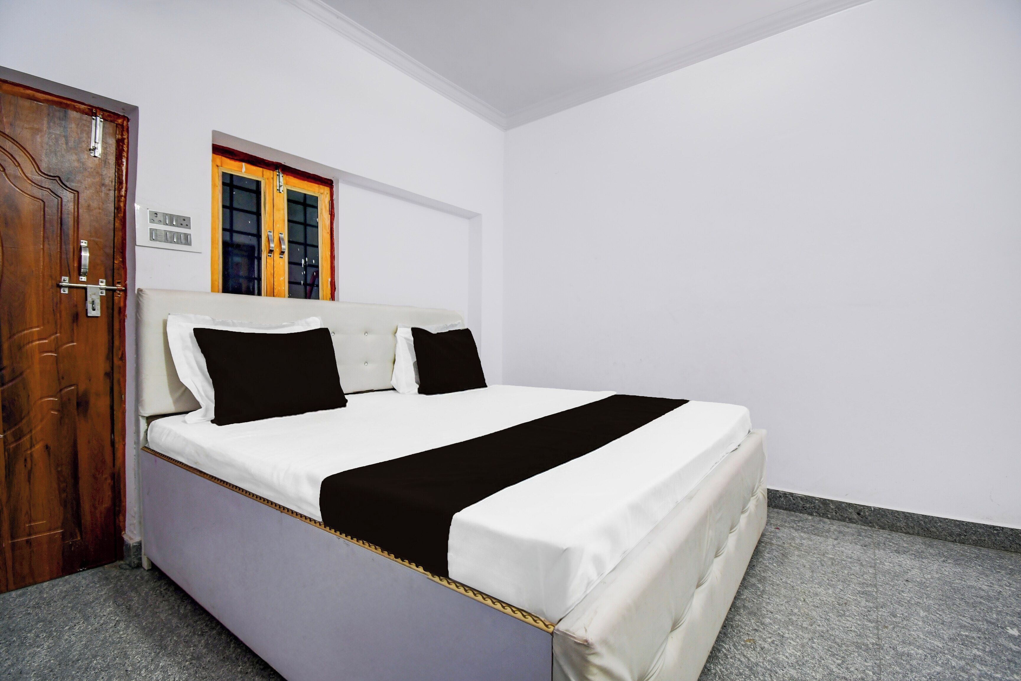 A single bed with a white mattress, two black pillows, and a white headboard in a room with white walls and a wooden door.