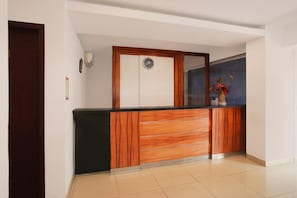 A reception desk with a wooden finish, a clock above it, and a vase with flowers on the counter. - Dancenter Hostel 77 Smart (Palembang)