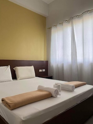 Hotellihuone, jossa on sänky, tyynyt, pääty, yöpöytä ja ikkuna, jossa on verhot.