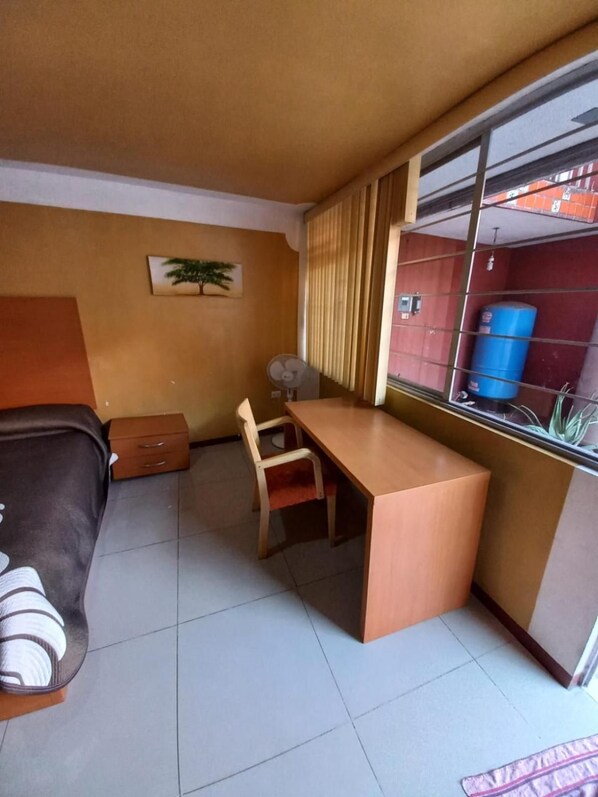 Une pièce de taille modeste comprenant un lit, un bureau, une chaise, un petit placard et une fenêtre avec des rideaux.