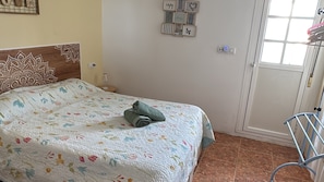 Ein Schlafzimmer mit einem Bett, einem Nachttisch, einem an der Wand befestigten Regal mit Bildern, einer Tür und einem Metallstuhl.