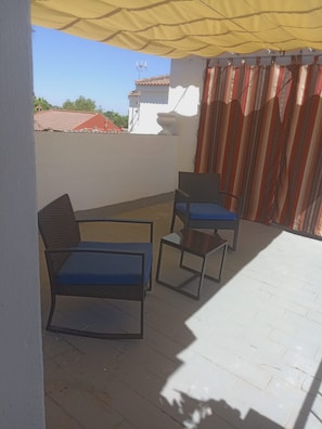 Ein überdachter Außenbereich mit zwei Stühlen, einem Tisch, einem Fliesenboden und Blick auf Gebäude im Hintergrund.