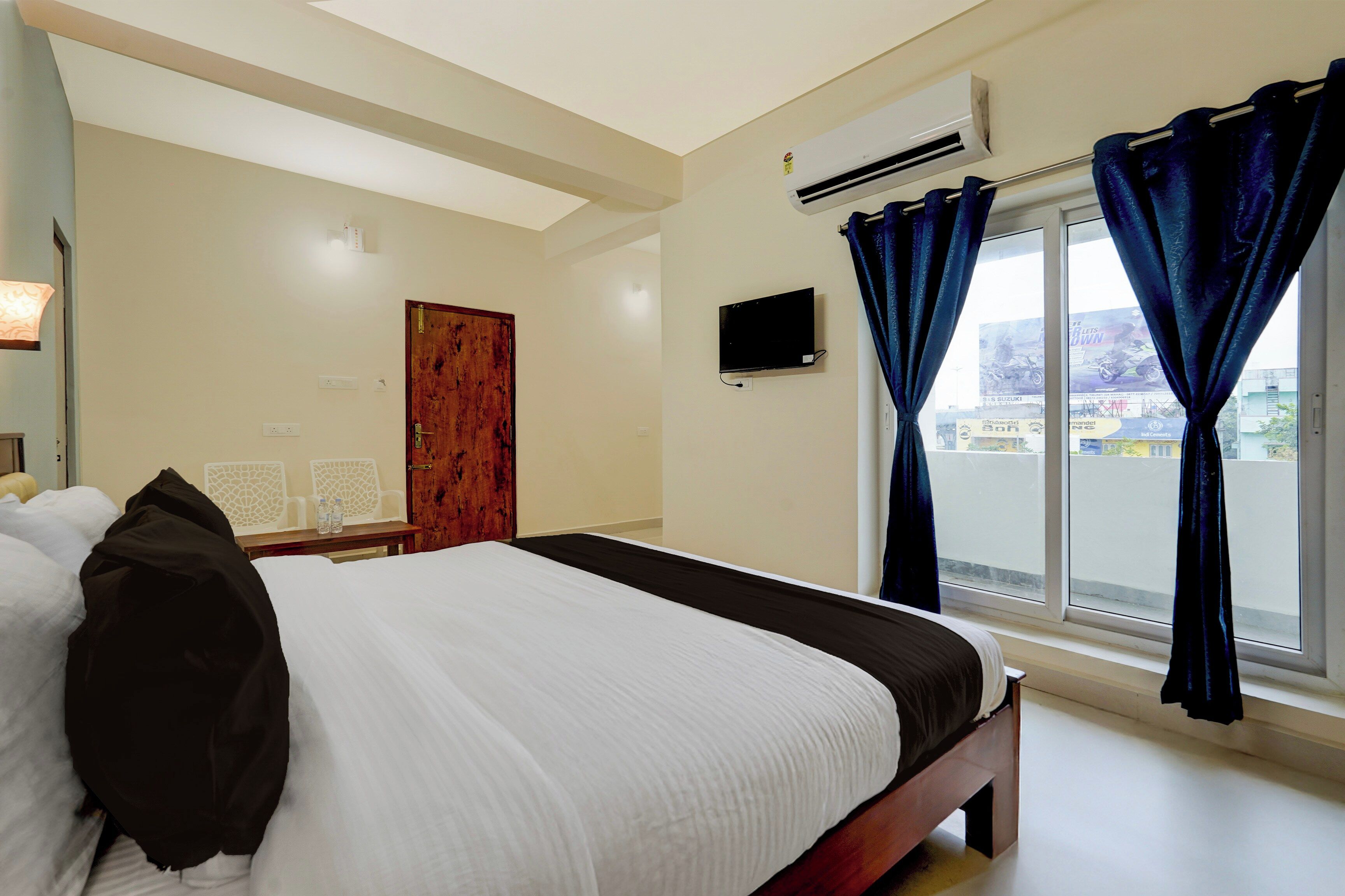 A hotel room with a bed, a flat-screen TV, a window with curtains, and an air conditioning unit.