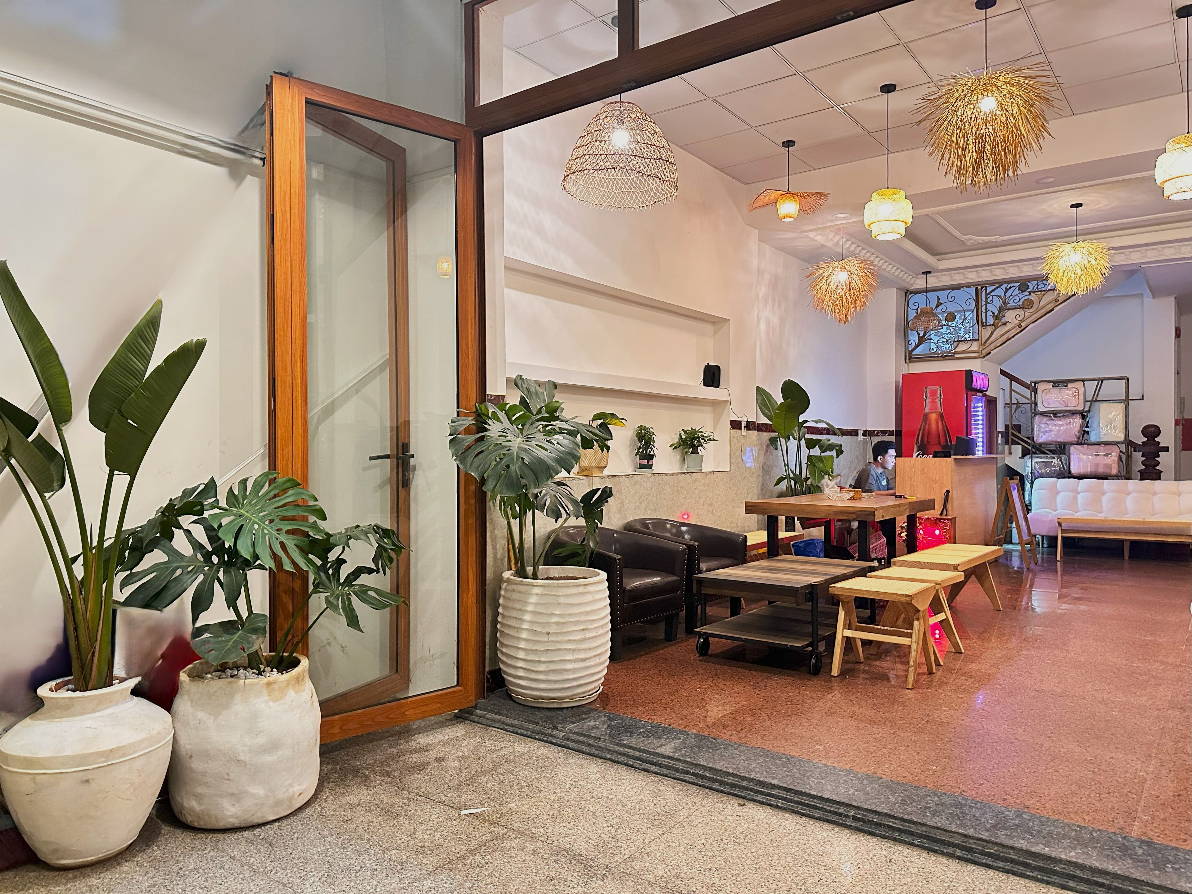 A hotel lobby with a wooden-framed door, potted plants, a seating area with a sofa and chairs, and hanging pendant lights.