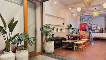 A hotel lobby with a wooden-framed door, potted plants, a seating area with a sofa and chairs, and hanging pendant lights.