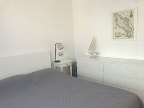 Een slaapkamer met een bed, een bureau, een stoel, een lamp, een klein model zeilboot en een kaart aan de muur.