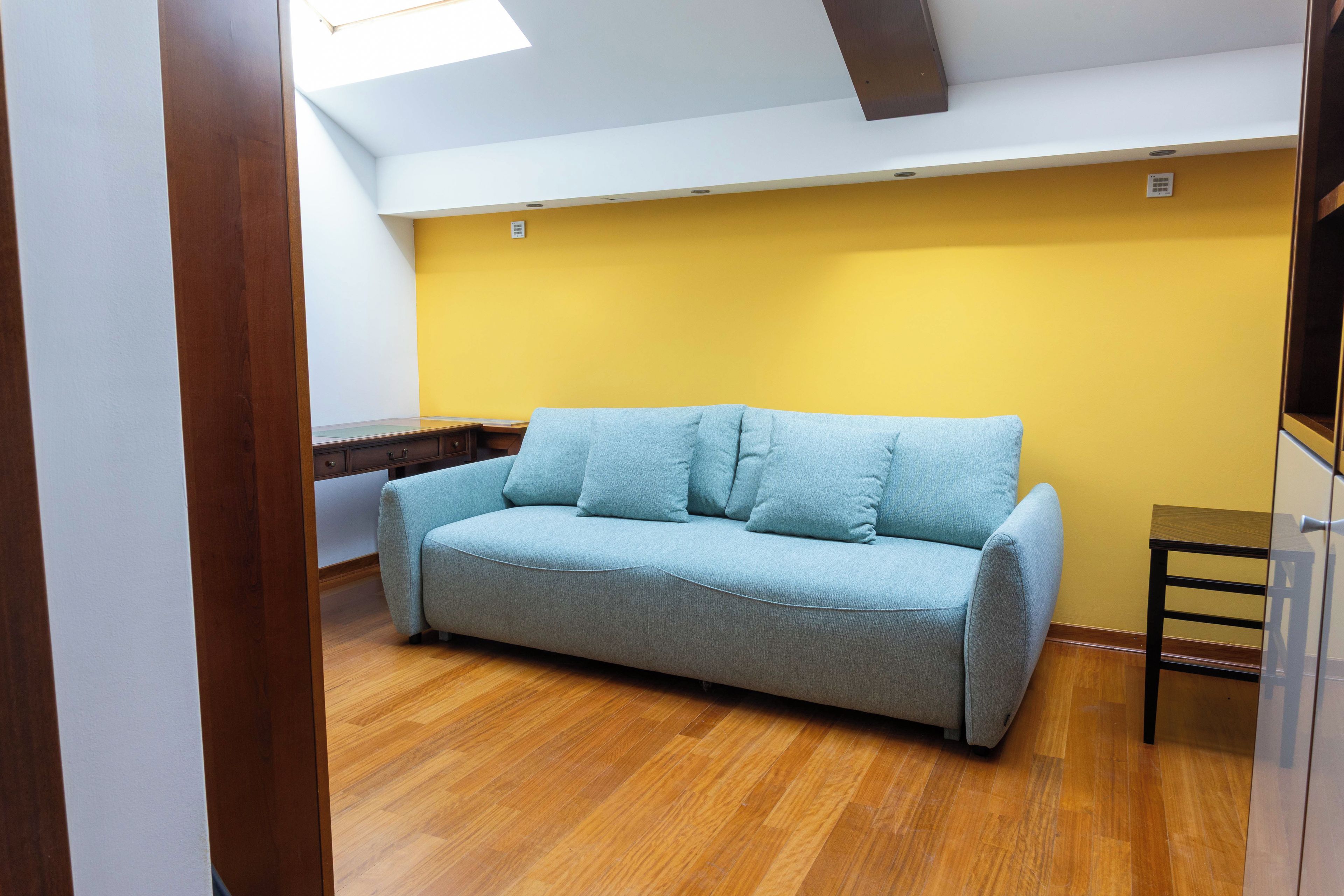 Ein Zimmer mit einem hellblauen Sofa, Holzboden, einer gelben Wand und einem Oberlicht.