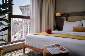 Ein Hotelzimmer mit einem großen Bett, einem Fenster mit Blick auf Gebäude, einem Nachttisch mit einem Buch und einem roten Dekorationselement.