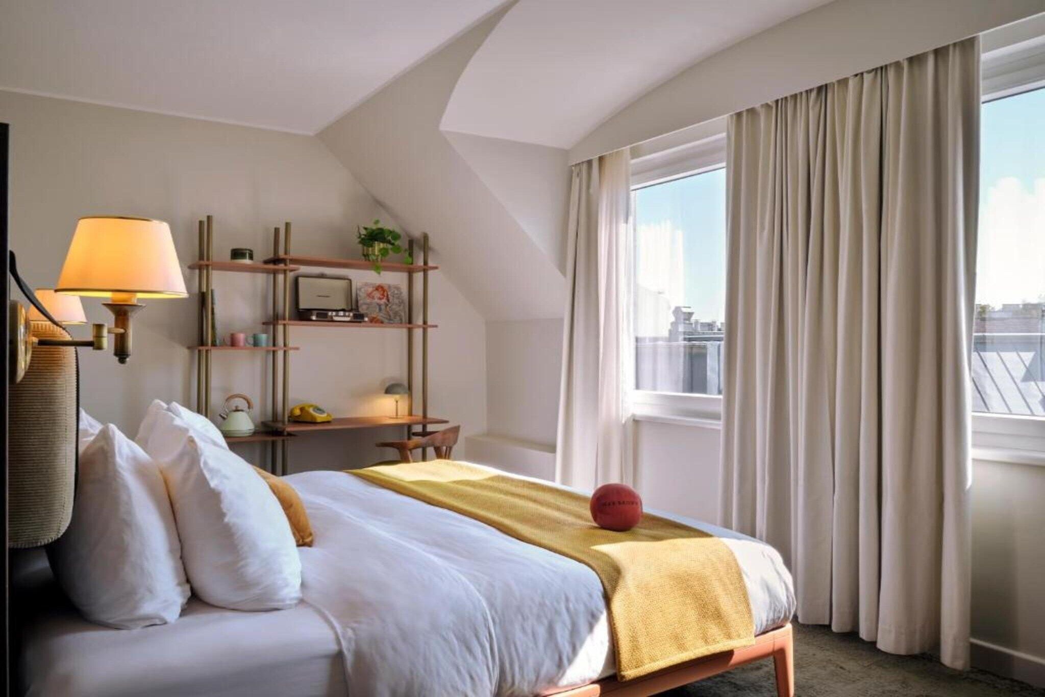 A hotel room with a bed, a desk, a lamp, and a window with curtains.